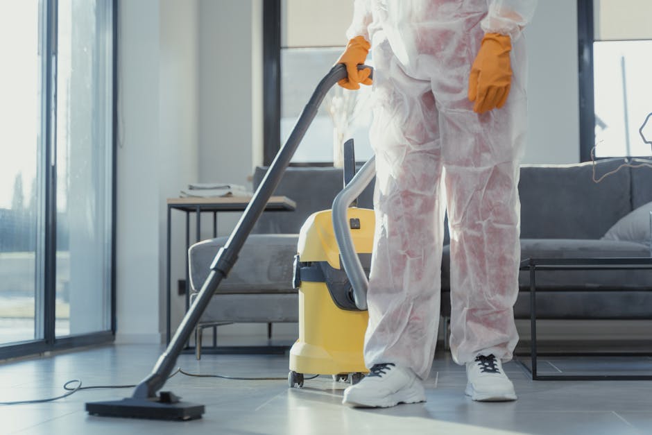 A professional cleaner from Deep Cleaning Bromley, dressed in a white protective suit and face mask, is performing deep sanitisation in a living room. The cleaner is using a fogging or fumigation devi